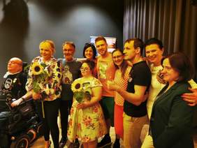 Pokaż zdjęcie: Liczna grupa osób pozuje do zdjęcia: kobiety i mężczyźni w różnym wieku, wśród nich są osoby z niepełnosprawnościami: mężczyzna na wózku inwalidzkim, kobieta z zespołem Downa oraz niewidomy mężczyzna. Wszyscy są uśmiechnięci. Dwie kobiety trzymają kwiaty.  W tle widać czarną kotarę.