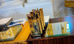 Pokaż zdjęcie: Na stole stoją statuetki w kolorze złotym o prostokątnym kształcie. Obok leżą dyplomy i szklane pamiątkowe tabliczki.