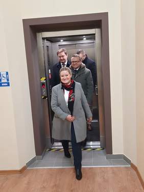 Pokaż zdjęcie: budynek Ośrodka Gminy na zdjęciu winda z której wysiadają osoby które przybyły na uroczyste otwarcie nowej windy w budynku w celu likwidacji barier dla mieszkańców ze szczególnymi potrzebami
