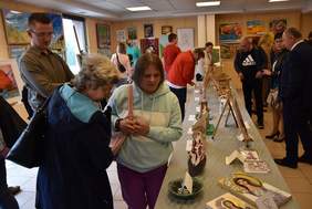 Pokaż zdjęcie: W dużej sali na ścianach są zawieszone obrazy, na środku stoją stoły z ustawionymi na nich rzeźbami. Kilkanaście osób chodzi i ogląda wystawione prace.