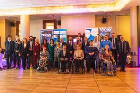 Pokaż zdjęcie: lebiscyt  Grupa ludzi, w tym osoby na wózkach inwalidzkich, pozują do wspólnego zdjęcia na tle banerów reklamowych i elegancko oświetlonego wnętrza. Ubrani są w eleganckie stroje - garnitury, sukienki i stylowe uniformy. W tle widać dwa duże banery z logo "TENZI" oraz inne oznaczenia, które sugerują, że wydarzenie ma charakter formalny lub związany ze wsparciem sportu bądź działalności integracyjnej. Sala jest przestronna, z błyszczącymi żyrandolami i nowoczesnym wystrojem.