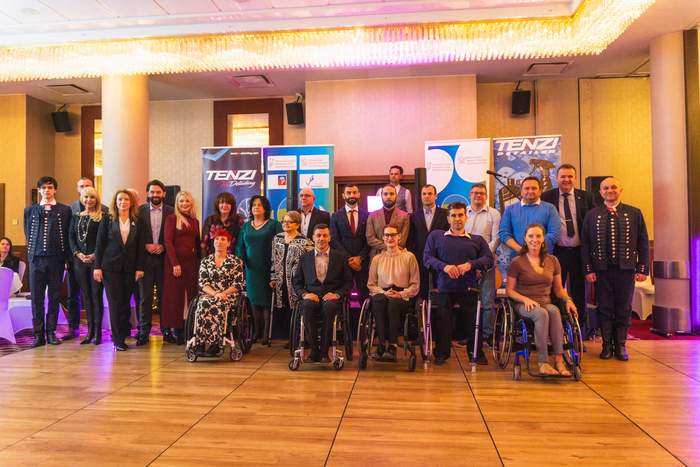 Pokaż zdjęcie: lebiscyt  Grupa ludzi, w tym osoby na wózkach inwalidzkich, pozują do wspólnego zdjęcia na tle banerów reklamowych i elegancko oświetlonego wnętrza. Ubrani są w eleganckie stroje - garnitury, sukienki i stylowe uniformy. W tle widać dwa duże banery z logo "TENZI" oraz inne oznaczenia, które sugerują, że wydarzenie ma charakter formalny lub związany ze wsparciem sportu bądź działalności integracyjnej. Sala jest przestronna, z błyszczącymi żyrandolami i nowoczesnym wystrojem.