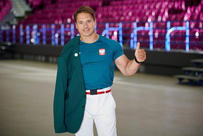 Pokaż zdjęcie: Mężczyzna w zielonej koszulce i białych spodniach, trzyma w ręku marynarkę, jest uśmiechnięty