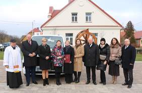 Pokaż zdjęcie: Grupa przedstawicieli samorządu, PFRON oraz instytucji lokalnych stojąca przed nowym pojazdem przystosowanym do przewozu osób z niepełnosprawnościami, na placu przed budynkiem Gminy Szumowo.