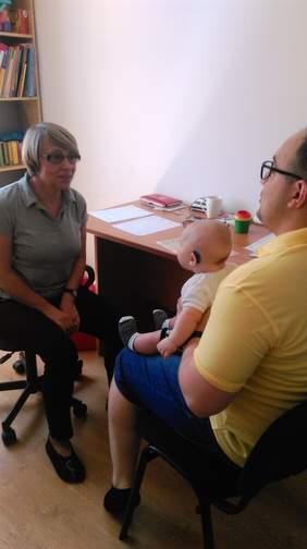 Pokaż zdjęcie: Kobieta i mężczyzna siedzący naprzeciwko siebie na krzesłach. Mężczyzna trzyma małego chłopca na kolanach.