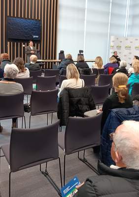 Pokaż zdjęcie: Na Sali konferencyjnej na podwyższeniu stoi kobieta przed nią na krzesłach siedzą ludzie którzy przysłuchują się jej wypowiedziom.