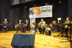 Występ sceniczny 4 mężczyzn i kobieta, siedzą z gitarami i przy instrumencie klawiszowym