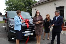 Pokaż zdjęcie: Cztery osoby uczestniczą w oficjalnym przekazaniu czarnego samochodu ozdobionego czerwoną kokardą i wstęgą. Grupa – trzy kobiety i mężczyzna w eleganckich strojach – stoi przed autem zaparkowanym na tle parterowego budynku i drzew.