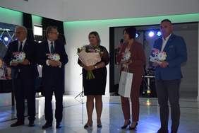 Pokaż zdjęcie: Pięć osób, dwie kobiety i trzech mężczyzn składa życzenia. Osoby te trzymają w ręku statuetki, kwiaty. 