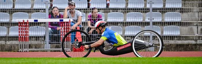 Pokaż zdjęcie: Na specjalnie przystosowanym rowerze w siedzisku jedzie mężczyzna. Ma na sobie sportowy strój kask i okulary.