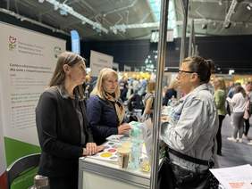 Pokaż zdjęcie: Trzy kobiety rozmawiają przy stoisku PFRON w hali sportowej; banery informacyjne projektu CIDON, na ladzie ulotki, woda i kolorowe naklejki. W tle tłum odwiedzających inne stoiska.