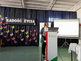 Pokaż zdjęcie: Przy mównicy stoi kobieta w zielonym garniturze.