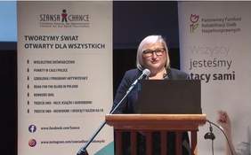 Pokaż zdjęcie: Kobieta z włosami koloru blond, nosząca okulary i ubrana w czarną marynarkę mówi do mikrofonu z tzw. ambony, na której stoi laptop. W tle widoczne są banery Fundacji Szansa dla Niewidomych oraz Państwowego Funduszu Rehabilitacji Osób Niepełnosprawnych.