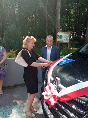 Pokaż zdjęcie: Kobieta w czarnej sukience z jasnymi włosami przecina biało czerwoną wstęgę na busie