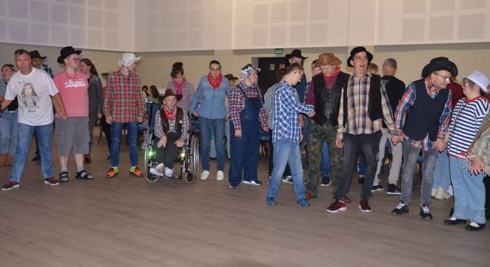 Pokaż zdjęcie: Grupa kilkunastu osób z niepełnosprawnością ubrana w większości w kraciaste koszule oraz mająca na głowie kapelusz. Osoby te podczas zabawy integracyjnej znajdują się w zadaszonym pomieszczeniu i stoją w półokręgu.