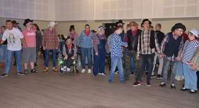 Pokaż zdjęcie: Grupa kilkunastu osób z niepełnosprawnością ubrana w większości w kraciaste koszule oraz mająca na głowie kapelusz. Osoby te podczas zabawy integracyjnej znajdują się w zadaszonym pomieszczeniu i stoją w półokręgu.