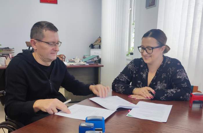 Pokaż zdjęcie: Przy stole w gabinecie mężczyzna i kobieta w okularach podpisują umowę. Na stole leżą pieczątki.