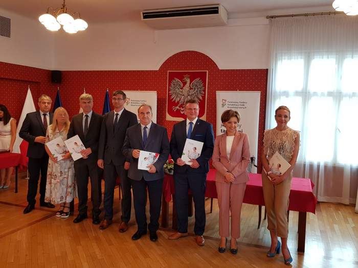 Pokaż zdjęcie: Wspólne zdjęcie przedstawicieli powiatów, osiem osób stojących obok siebie w sali konferencyjnej na tle godła. niektóre osoby trzymają w rekach teczki z podpisanymi dokumentami