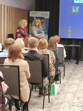 Dwie kobiety w kolorowych bluzkach, stoją, grupa osób siedzi