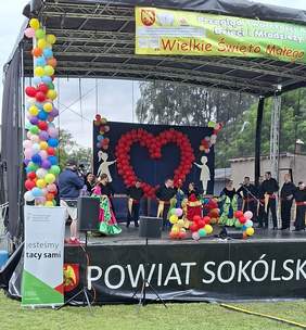 Pokaż zdjęcie: Scena plenerowa udekorowana kolorowymi balonami, z dużym sercem z czerwonych balonów na tle niebieskim. Na scenie dzieci i młodzież w kolorowych strojach ludowych wykonują taniec. W tle baner wydarzenia "Wielkie Święto Małego Człowieka". Po lewej stronie stoi baner z napisem „Wszyscy jesteśmy tacy sami”, promujący integrację społeczną. Na dole widoczny napis „Powiat Sokólski”.