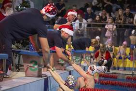 Pokaż zdjęcie: Grupa trenerów w czapkach Mikołaja pomaga młodym pływakom podczas zawodów na basenie. W tle widoczna jest publiczność oraz dzieci w strojach pływackich.