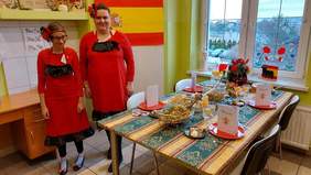 Pokaż zdjęcie: Przy stole stoją dwie kobiety ubrane w czerwone suknie z czarnymi wstawkami. We włosach mają wpleciony czerwony kwiat. Stół nakryty obrusem. Na nim potrawy, napoje sztućce i czerwone talerze. Na talerzach stoi karka z menu. Obok flaga hiszpańska z 