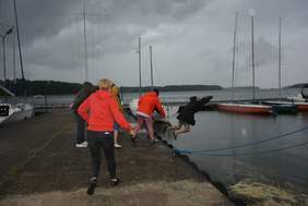 Pokaż zdjęcie: Mężczyznę w ubraniu dwie osoby wrzucają do jeziora. Oprócz wrzucających na pomoście idzie kilka osób w kurtkach. W tle żaglówki. Na jeziorze widać odznaczające się krople deszczu.