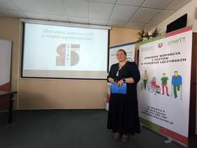 Pokaż zdjęcie: Kobieta w długiej ciemnej sukience prowadzi wykład.