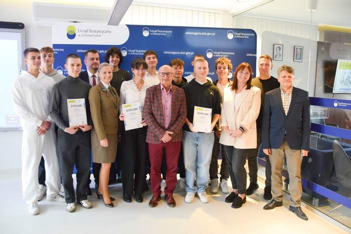 Pokaż zdjęcie: Zwycięska młodzież wraz z opiekunami oraz przedstawicielami instytucji, pozuje do pamiątkowej fotografii. Kilka osób trzyma w dłoniach dyplomy. Grupa stoi na tle ścianki promocyjnej Urzędu Statystycznego w Zielonej Górze.