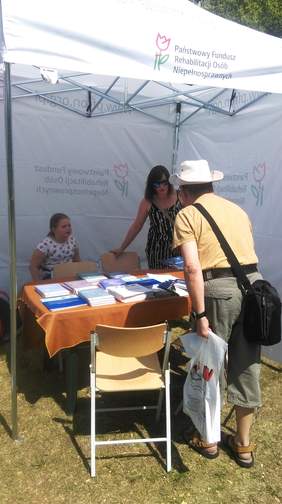 Pokaż zdjęcie: Kobieta stojąca przy stoliku z materiałami informacyjnymi podająca mężczyźnie ulotki, w tle siedzi na krzesełku dziecko –Warszawa