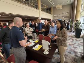 Pokaż zdjęcie: Dwóch mężczyzn w ciemnych koszulach rozmawia z grupą osób przy stoliku, na którym leżą broszury i materiały informacyjne.