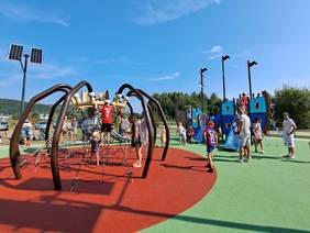 Pokaż zdjęcie: Piękna pogoda, bezchmurne niebo, na macie bezpiecznej stoi wielki pająk z którego wychodzą liny ułożone w siatkę. Obok stoi duży statek z trzema masztami. Po placu i zabawkach biegają dzieci.