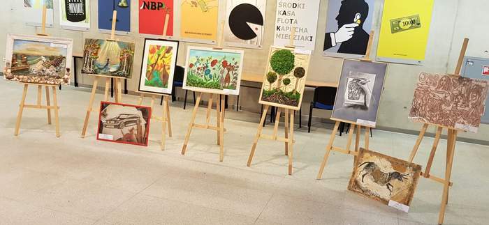 Pokaż zdjęcie: Na siedmiu sztalugach ustawione zostały prace wielkopolskich laureatów XIX edycji Konkursu Plastycznego „Sztuka Osób Niepełnosprawnych”. Zostały one wykonane w pięciu kategoriach, czyli: rzeźba kameralna i płaskorzeźba, malarstwo i witraż, tkanina i aplikacja, rysunek i grafika oraz fotografia. W tle widocznych jest dziewięć wielobarwnych plakatów zawieszonych na ścianie budynku Centrum Edukacyjnego Usług Elektronicznych Uniwersytetu Ekonomicznego w Poznaniu. Niniejsze banery prezentują różne zagadnienia związane z ekonomią.
