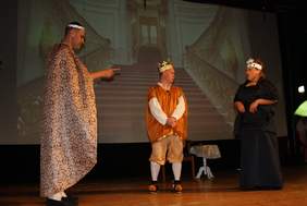 Pokaż zdjęcie: Na scenie stoi dwóch mężczyzn i kobieta. Kobieta jest ubrana w długą czarną suknię na głowie ma srebrną koronę, mówi do mężczyzn, którzy na nią patrzą. Jeden z nich  jest ubrany w pomarańczowy ozdobny strój i ma na głowie złotą koron