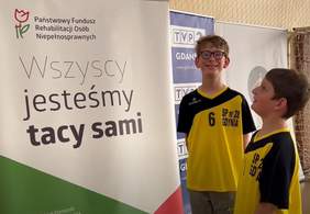 Pokaż zdjęcie: Dwóch chłopców w sportowych koszulkach stoi przed banerem z logo PFRON i z napisem „Wszyscy jesteśmy tacy sami” jeden z nich w okularach śmieje się do obiektywu aparatu.
