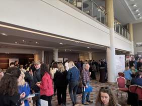 Pokaż zdjęcie: Grupa ludzi rozmawia i stoi przy stoiskach informacyjnych