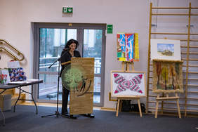 Pokaż zdjęcie: Kobieta stojąca przy mównicy wygłasza przemówienie podczas gali konkursowej. Obok niej znajdują się kolorowe prace plastyczne ustawione na sztalugach, a po lewej stronie widoczny jest stół z nagrodami.