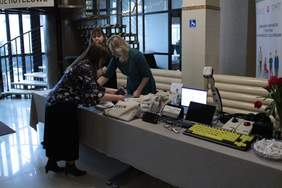 Pokaż zdjęcie: Recepcja hotelu w którym stoi długi stół przykryty beżowym obrusem. Przy stole stoją trzy kobiety i układają materiały reklamowe. Po prawej stronie stołu leżą klawiatury oraz urządzenia wspomagające komunikację