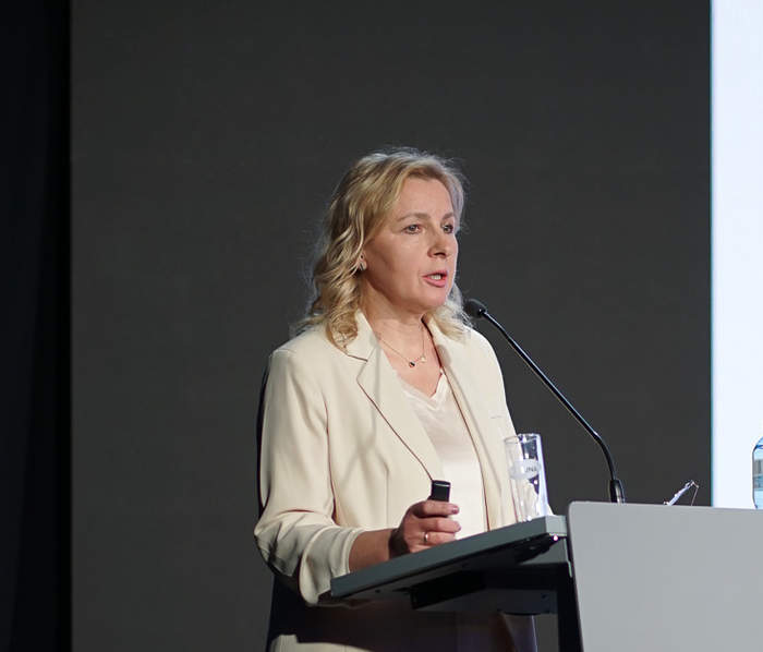 Pokaż zdjęcie: Przy mównicy stoi kobieta blondynka w białej marynarce. W tle widać slajd z prezentacji.