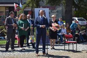 Jasny słoneczny dzień, na rynku na tle wystawy ze zdjęciami prac stoi kilka osób, mężczyzna przemawia, w tyle widać innych