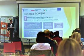 Po lewej strony stoi prelegentka, a obo knie j ekran, na którym wyświetlany jest slajd zatytuowany "Zadania SCWEW