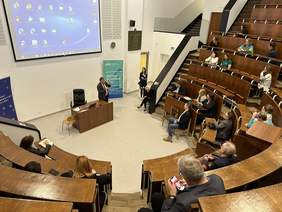 Pokaż zdjęcie: Na auli ułożonej w okrąg siedzą ludzie. Na samym dole na środku stoi mężczyzna w marynarce i, koszuli i krawacie trzyma mikrofon. Za nim duży ekran i dwa banery reklamowe.