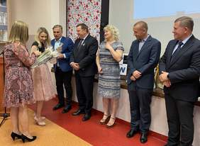 Pokaż zdjęcie: W rzędzie stoi czterech mężczyzn i kobieta. Wszyscy mężczyźni ubrani w garniturach. Jeden z nich Odbiera statuetkę od kobiety stojącej naprzeciwko. W tle stoi mównica i szafka z trofeami.