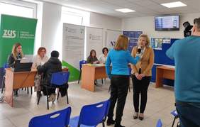 Pokaż zdjęcie: Sala Obsługi Klientów w NFZ Lublin. w tle widać stoliki przy których siedzą pracownicy PFRON, ZUS i KRUS. Kobieta blondynka w beżowej marynarce udziela wywiadu Pani redaktor ubranej w niebieską marynarkę. Po prawej stronie widać mężczyznę filmującego wywiad.