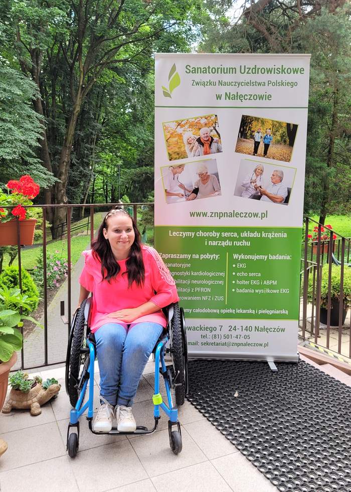 Pokaż zdjęcie: Pani Weronika - młoda uśmiechnięta kobieta na wózku inwalidzkim. Za panią Weroniką rollup z ORK w Nałęczowie