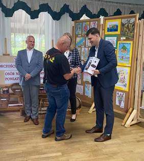 Pokaż zdjęcie: Uśmiechnięty mężczyzna wręcza nagrodę laureatowi. Obok kobieta i mężczyzna. Za nimi prace konkursowe