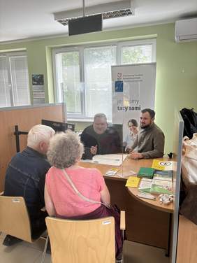 Pokaż zdjęcie: racownicy Państwowego Funduszu Rehabilitacji Osób Niepełnosprawnych prowadzą dyżur informacyjny. Przy stole siedzą dwaj pracownicy oraz kobieta zainteresowana uzyskaniem informacji. Na biurku znajdują się materiały informacyjne i ulotki PFRON.