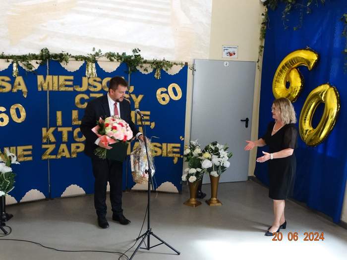 Pokaż zdjęcie: Dyrektor Oddziału wręcza kwiaty Dyrektor Ośrodka, oboje są ubrani na galowo, na tle ścianki z napisem 60-lecie