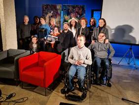 Pokaż zdjęcie: Uczestnicy spotkania pozują do zdjęcia na scenie. Wśród uczestników są kobiety, mężczyźni, osoby na wózkach oraz niewidome.