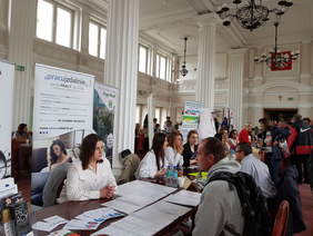 Sala z kolumnami, zebrami podczas giełdy, część osób siedzi za stołami, inni stoją, rozmawiają